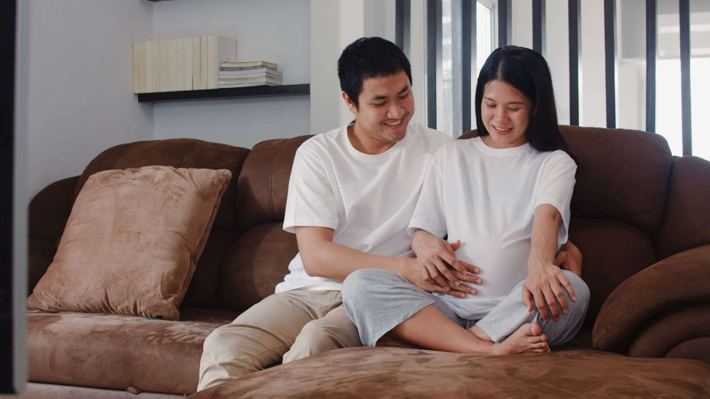 Pasangan Asia duduk di sofa sedang menyentuh perut istri yang sedang hamil.