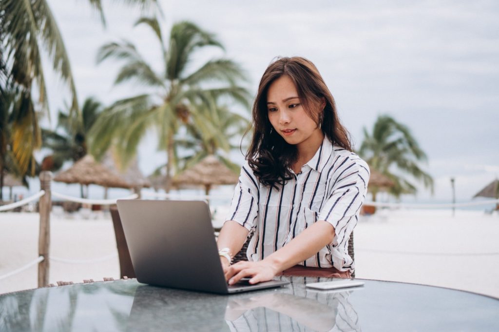 Seorang wanita Asia sedang bekerja menggunakan laptop saat di pantai.