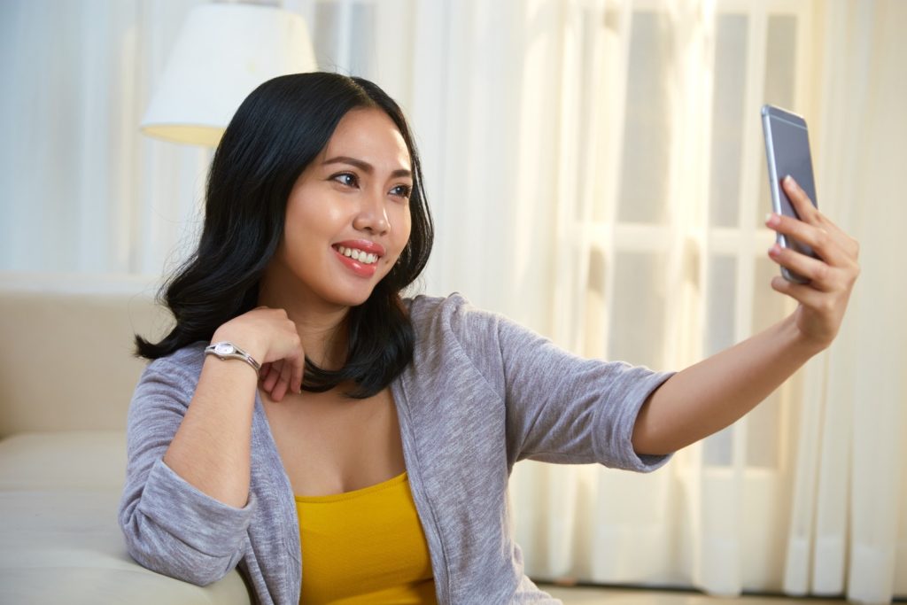 Seorang wanita Asia sedang selfie menggunakan smartphone miliknya.