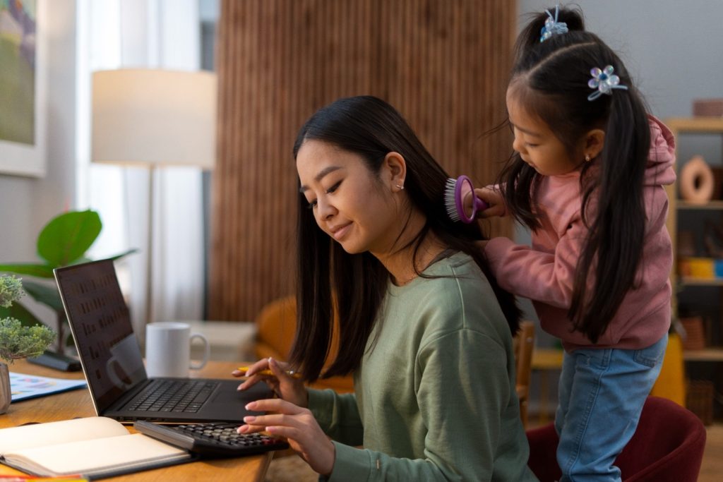 Seorang anak menyisir rambut ibunya yang sedang bekerja dari rumah menggunakan laptop.