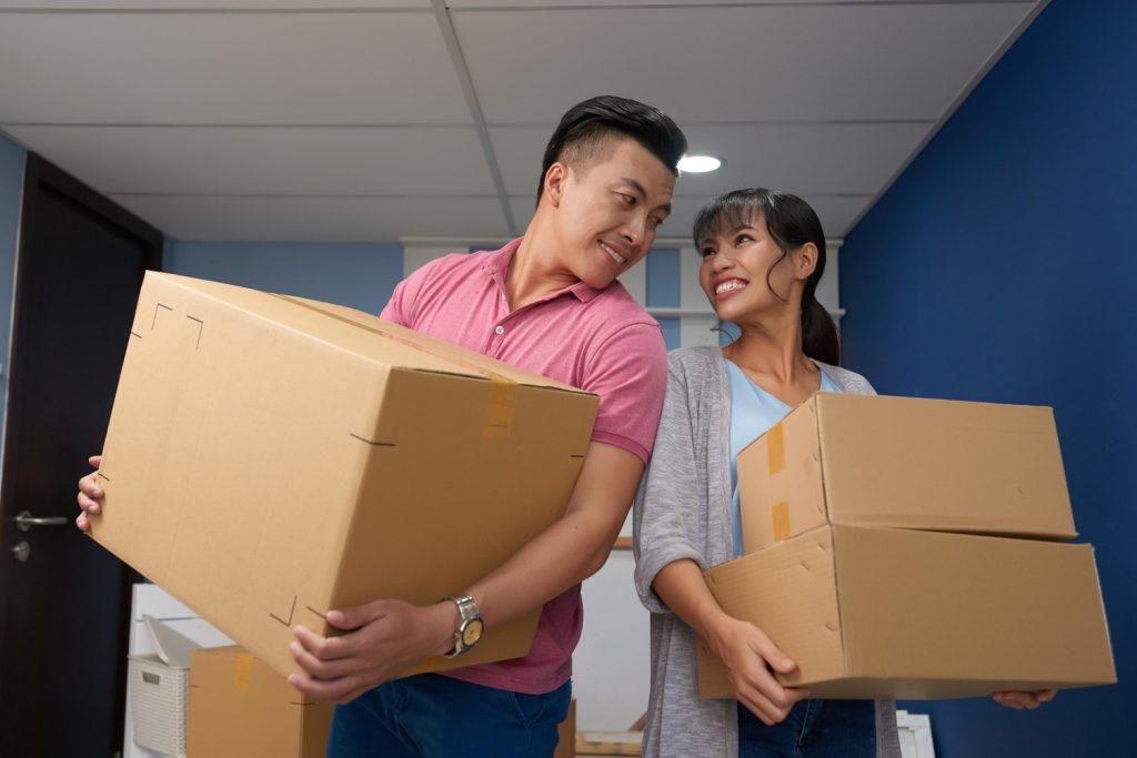 Sepasang suami istri memegang kardus untuk pindahan rumah.