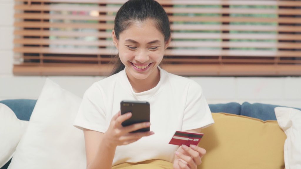 Seorang wanita tersenyum melihat layar ponsel sambil memegang kartu kredit.
