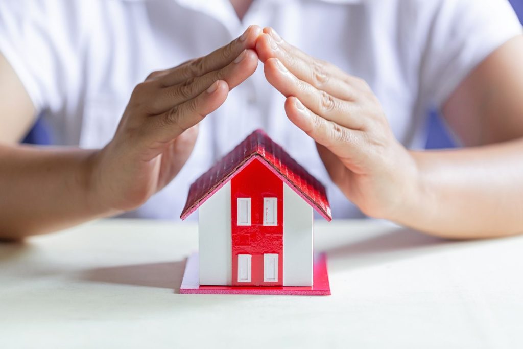 Potret sepasang tangan menutup figurin miniatur rumah.